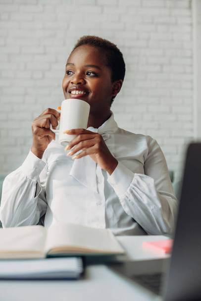 Young african woman looking away dreaming about weekends while drinking coffee in morning at office. Enjoying drinking coffee sitting in cozy modern office. - Zdjęcie, obraz