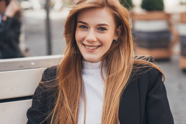 Businesswoman portrait with long foxy hair and freckled face looking at smiling at camera, sitting on bench in front the office building. Positive person - 写真・画像