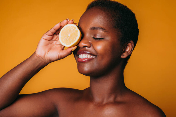 Smiling afro woman covering one eye with lemon halves isolated over orange background. Beauty, skin care and detox concept. - Foto, immagini