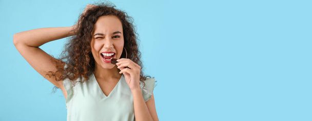Beautiful young African-American woman eating delicious chocolate candy on blue background with space for text - Zdjęcie, obraz