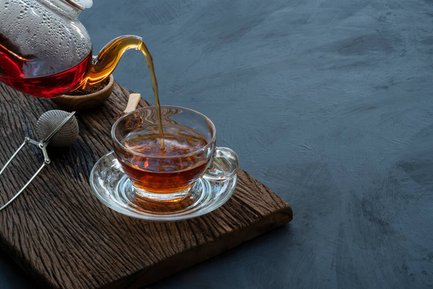 Process brewing tea, Hot tea water is poured from the teapot into a cup on the old wood plank with dark kitchen table background, still life relaxing time dark mood style. - Photo, Image