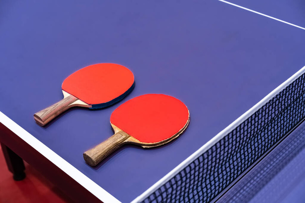 Два настільних тенісних ракетки для гри покладені поруч із сіткою на синьому столі. Це одне спортивне обладнання для пінг-понг для гравців
 - Фото, зображення