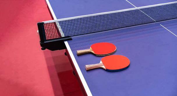 Два настільних тенісних ракетки для гри покладені поруч із сіткою на синьому столі. Це одне спортивне обладнання для пінг-понг для гравців
 - Фото, зображення