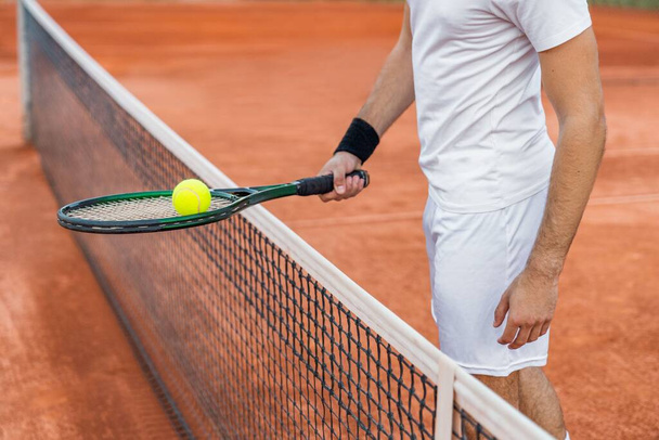 Junger Mann hält den Schläger mit dem Ball in der Nähe des Netzes auf einem unbefestigten Tennisplatz. - Foto, Bild
