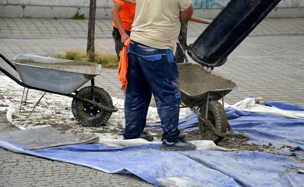 Men in work clothes, midre colors. concrete mixer. truck chute trough, pouring, concrete into wheelbarow. bricklayers drive to the construction site. tarpaulin covers the pavemen - Photo, image