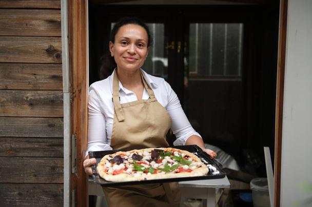 Pleasant multiethnic woman, housewife in a beige chefs apron, smiling cutely looking at camera, holding raw pizza pan before putting it in the oven, standing in the entrance of rustic summer kitchen - Foto, Bild
