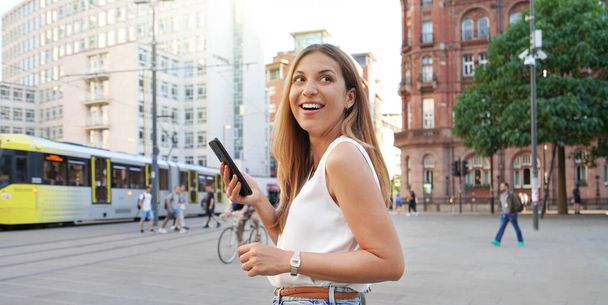 Attractive laughing woman holding smartphone walking on urban background. Looking to the side. Banner cropped view. - Fotó, kép