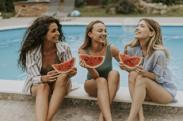 Three cute young women sitting on by the swimming pool and eating watermelon in the house backyard - Foto, Bild