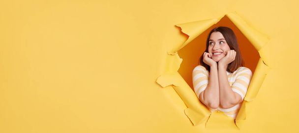 Smiling delighted woman wearing striped t shirt, expreressing positve emotions, copy space for advertisement or promotional text, posing standing through yellow paper torn hole. - Foto, imagen