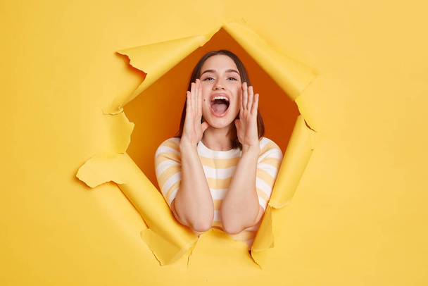 Horizontal shot of positive woman wearing striped t shirt posing standing through yellow paper torn hole, screaming, announcing advertisement with optimistic expression. - Foto, Imagen