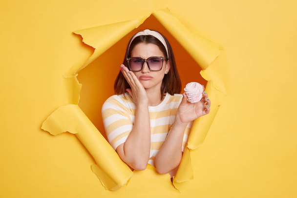 Portrait of unhealthy sick woman wearing hair band and striped t shirt standing in yellow paper hole, feeling pain after eating sugary dessert, touching her cheek. - Foto, Imagen