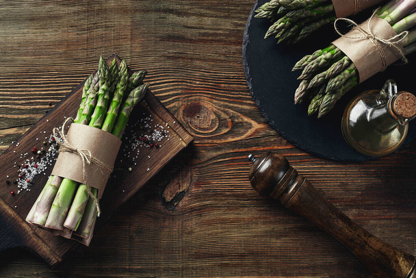 Bunches of an edible, uncooked stems of asparagus on wooden background. Fresh, green vegetables with olive oil and seasonings, kitchen pestle, top view. Healthy eating. Fall harvest, agricultural - Fotoğraf, Görsel
