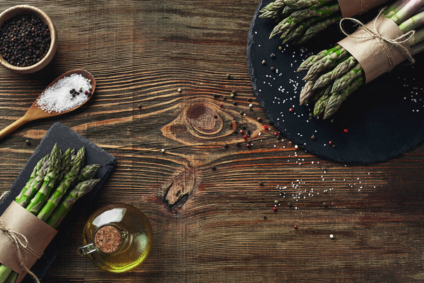 Bunches of an edible, fresh stalks of asparagus on wooden background. Green vegetables with olive oil and seasonings, top view. Healthy meal. Fall harvest, agricultural farming concept. - Foto, Imagen