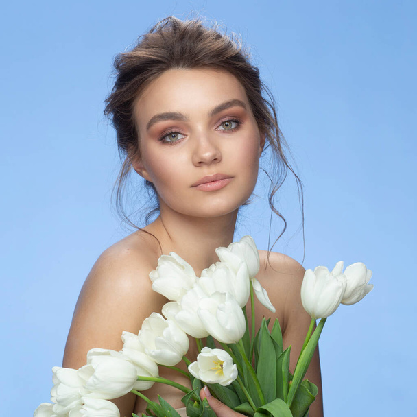 Close up portrait of the young woman with natural makeup and hairstyle looking at the camera, white tulips covered half of female's face, isolated over blue. - Photo, image