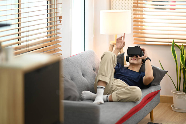 Young man in casual clothes lying on couch and watching movie or play video game on virtual reality headset. - Foto, Imagem