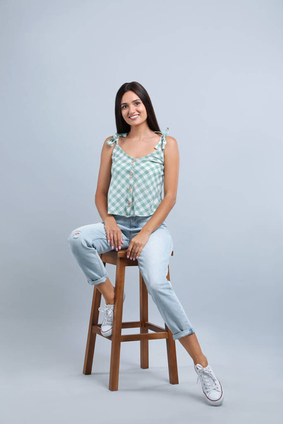Beautiful young woman sitting on stool against light grey background - Fotografie, Obrázek