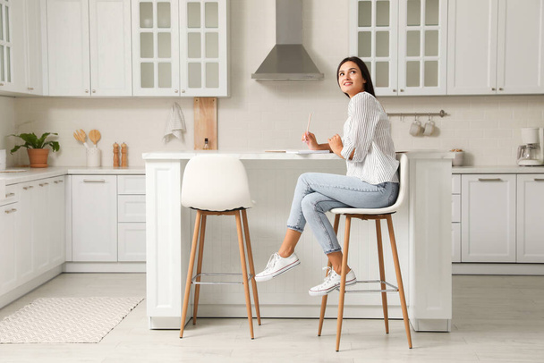 Beautiful young woman with notebook sitting on stool in kitchen - Zdjęcie, obraz