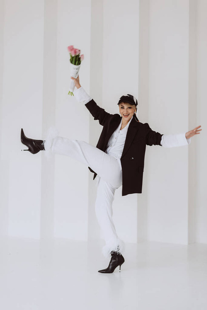 A happy stylish woman in a fashionable outfit, a light white suit with feathers, a black jacket, a baseball cap and with a bouquet of flowers in her hands is having fun. Soft selective focus. - Φωτογραφία, εικόνα