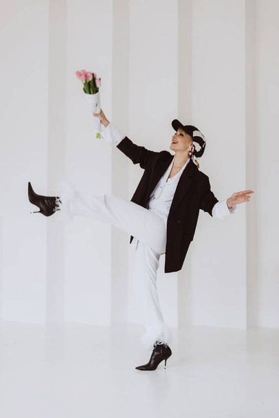 A happy stylish woman in a fashionable outfit, a light white suit with feathers, a black jacket, a baseball cap and with a bouquet of flowers in her hands is having fun. Soft selective focus. - Fotoğraf, Görsel