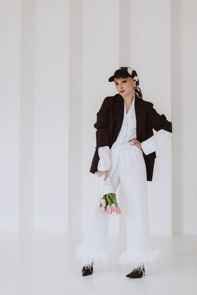 A young stylish woman in a fashionable outfit, a light white suit with feathers, a black jacket, a baseball cap and a bouquet of flowers in her hands poses on a white background. Soft selective focus. - Photo, Image