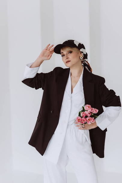 A beautiful woman in a fashionable outfit, a light white suit and a black jacket, a baseball cap and with a bouquet of tulips in her hands is happy. Soft selective focus. - Фото, изображение