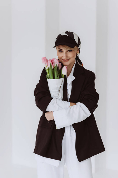A beautiful woman in a fashionable outfit, a light white suit and a black jacket, a baseball cap and with a bouquet of tulips in her hands is happy. Soft selective focus. - Foto, Imagem