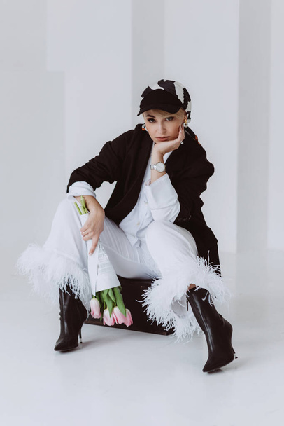 A stylish young woman in a light suit with feathers and a black jacket poses while sitting with a bouquet of tulips in her hands. Modern fashion. Soft selective focus. - Zdjęcie, obraz