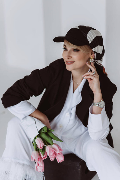 A stylish young woman in a light suit with feathers and a black jacket poses while sitting with a bouquet of tulips in her hands. Modern fashion. Soft selective focus. - Foto, Imagen