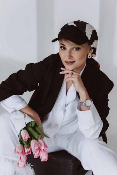 A stylish young woman in a light suit with feathers and a black jacket poses while sitting with a bouquet of tulips in her hands. Modern fashion. Soft selective focus. - Foto, Imagem