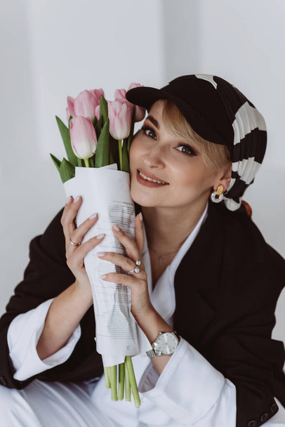 A stylish young woman in a light suit with feathers and a black jacket poses while sitting with a bouquet of tulips in her hands. Modern fashion. Soft selective focus. - Foto, immagini