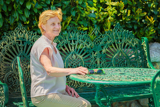 Mature attractive woman traveler sitting alone on the terrace of coffee shop in beautiful park in Bulgaria. Active life of the elderly in retirement, active seniors - Φωτογραφία, εικόνα