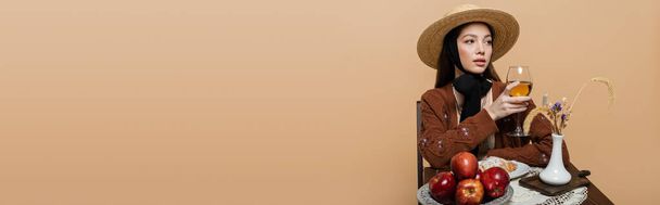 Stylish woman in sun hat holding glass of wine near food on table isolated on beige, banner  - Foto, Imagem