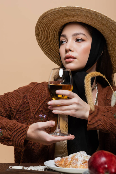 Stylish woman in straw hat posing with glass of wine near croissant on table isolated on beige  - Photo, Image