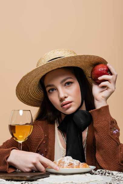 Young model in straw hat holding apple and looking at camera near croissant and wine isolated on beige  - Valokuva, kuva