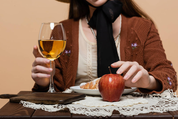 Обрізаний вид розмитої жінки торкається келиха вина та яблука на столі ізольовано на бежевому
  - Фото, зображення