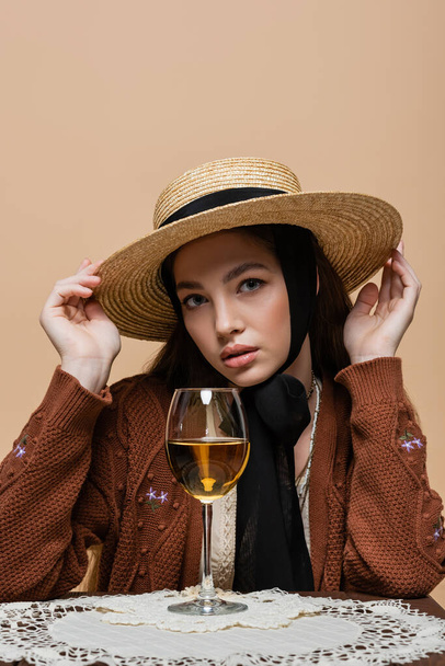 Trendy woman in straw hat looking at camera near glass of wine on table isolated on beige  - Photo, Image