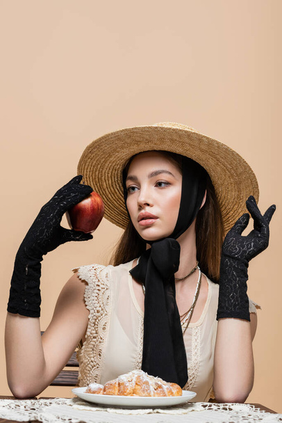 Trendy woman in straw hat and gloves holding apple near croissant isolated on beige  - Foto, Bild