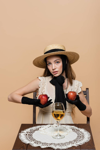 Fashionable woman in gloves and sun hat holding apples near wine isolated on beige  - Фото, зображення
