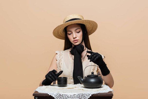 Stylish woman in straw hat and gloves sitting near teapot and cup isolated on beige  - Фото, изображение