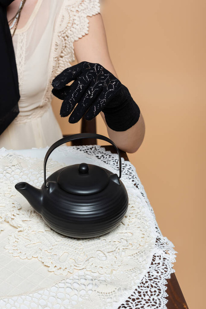 Cropped view of stylish woman in glove sitting near teapot on table isolated on beige  - 写真・画像
