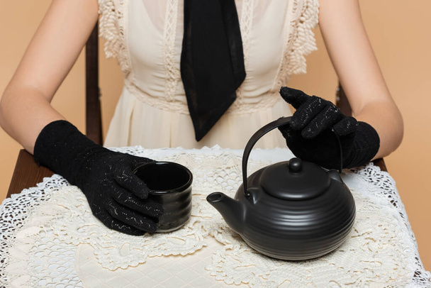 Обрізаний вид жінки в рукавичках тримає чашку і чайник на столі ізольовані на бежевому
  - Фото, зображення