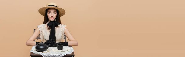 Fashionable woman in straw hat and gloves looking at camera near black teapot on table isolated on beige, banner  - 写真・画像