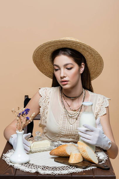 Trendy model in sun hat holding milk and butter near bread on table isolated on beige  - Foto, immagini