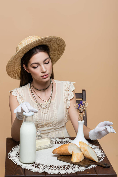 Pretty woman in sun hat and gloves sitting near bread and milk on table isolated on beige  - Fotografie, Obrázek