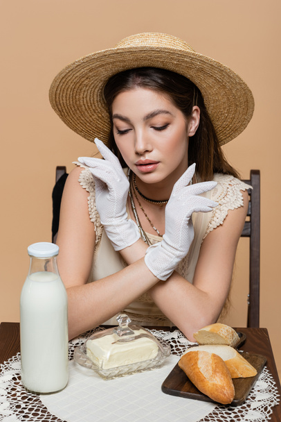 Brunette woman in straw hat and gloves looking at food and milk on table isolated on beige  - Fotografie, Obrázek