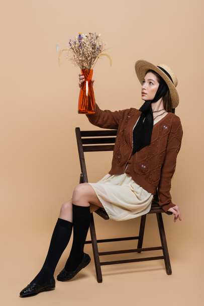 Stylish woman in cardigan and sun hat holding plants in vase on beige background  - Fotoğraf, Görsel