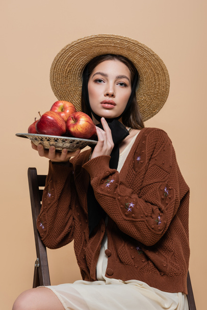 Portrait of stylish woman in straw hat holding apples on plate on beige background  - Zdjęcie, obraz
