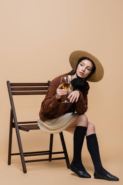 Trendy young woman in straw hat holding wine while sitting on chair on beige background  - Foto, Imagen