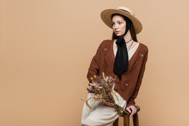 Brunette woman in straw hat holding dry flowers near chair isolated on beige  - Foto, Imagem