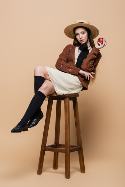 Stylish woman in straw hat holding apple while sitting on chair on beige background  - Fotografie, Obrázek